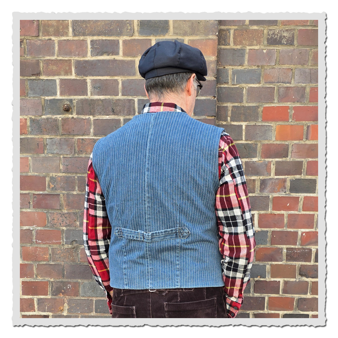 Rückansicht eines Mannes mit Schiebermütze, kariertem Hemd und blauer Weste vor Backsteinwand.